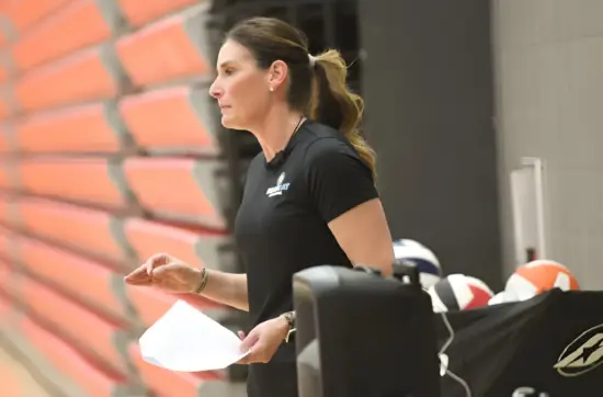 Side profile of Cathy Helin reviewing notes near volleyball equipment, showcasing coaching and athlete development strategies.