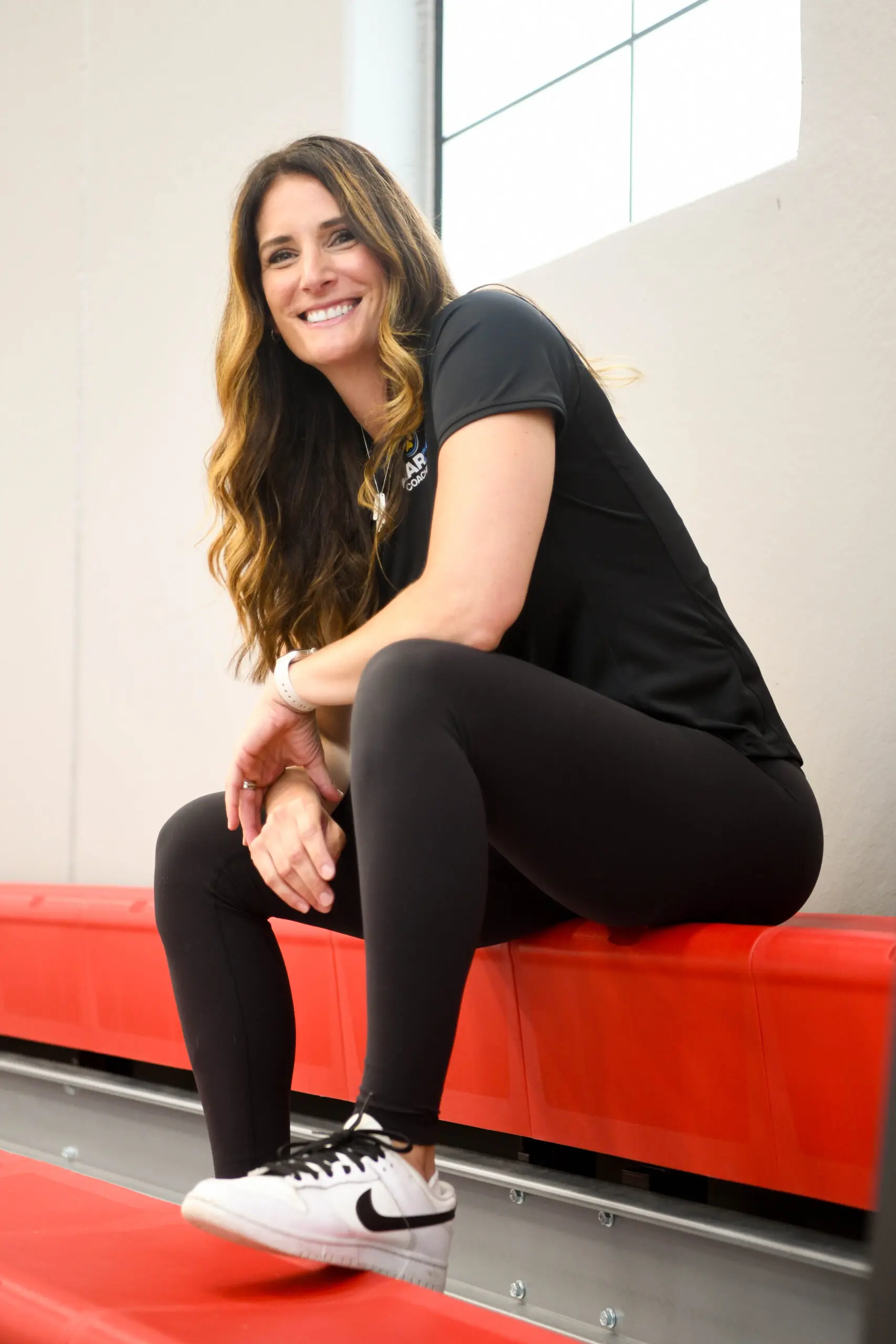 Cathy Helin crouches on a red gym bench, smiling an coaching style focused on athlete growth.