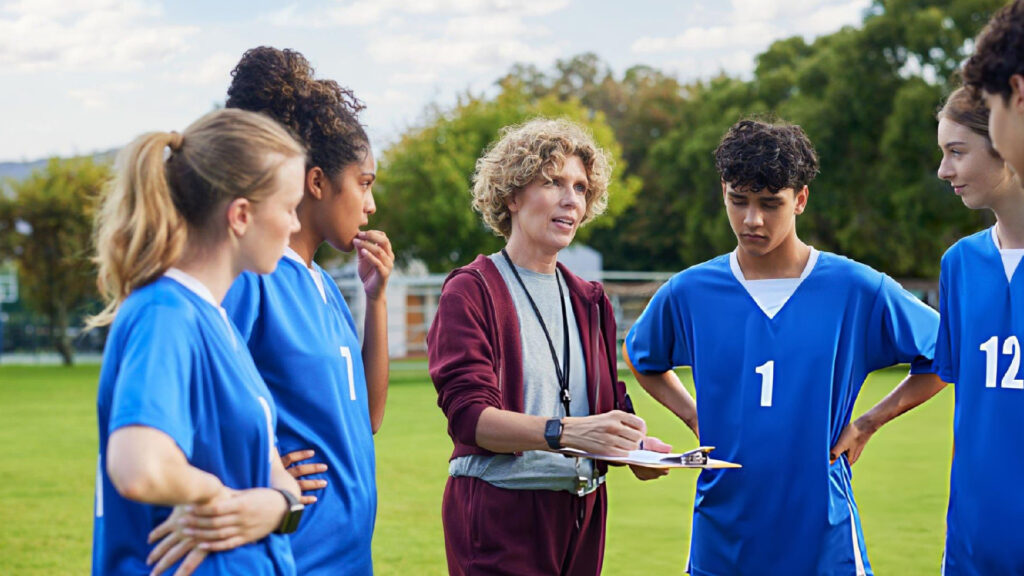 Sports coach guiding young athletes during team discussion, developing leadership, communication, teamwork, confidence, and performance on field.