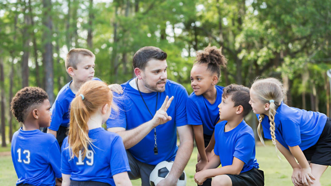 Coach kneels with young soccer players, giving team instructions before practice outdoors while everyone listens attentively together today.