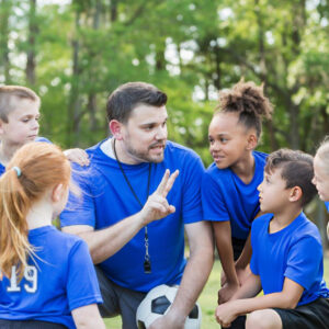 Coach kneels with young soccer players, giving team instructions before practice outdoors while everyone listens attentively together today.