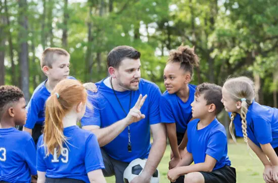 Coach kneels with young soccer players, giving team instructions before practice outdoors while everyone listens attentively together today.