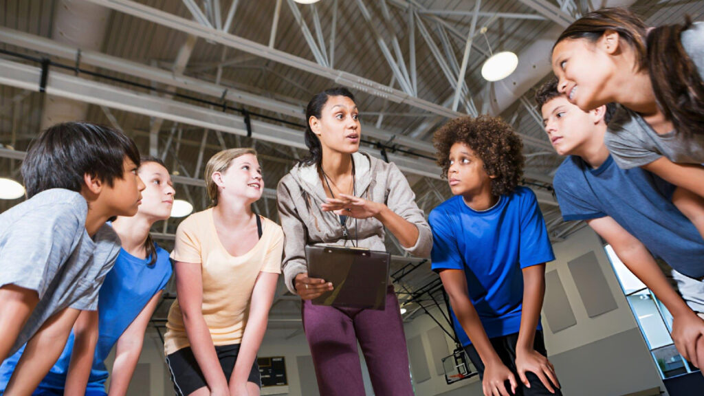 Coach explains game strategy to youth athletes during indoor practice as focused teammates gather closely and listen carefully.