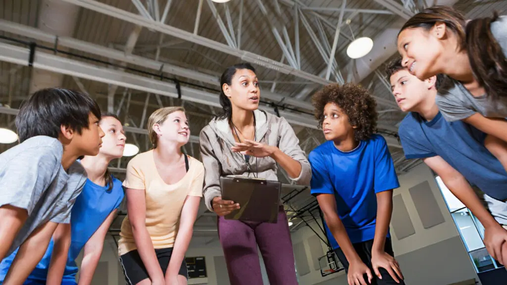 Coach explains game strategy to youth athletes during indoor practice as focused teammates gather closely and listen carefully.