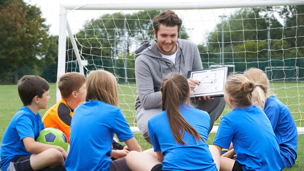 Soccer coach uses tactics board, teaching young players strategy before match while team listens attentively on field.