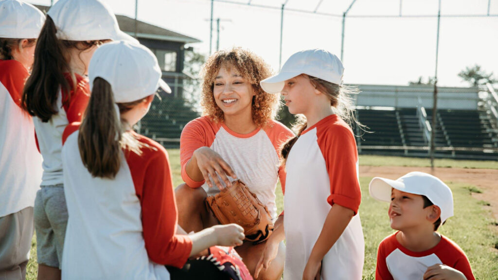 Young girls in baseball uniforms gather outdoors with a coach during a friendly practice session together.