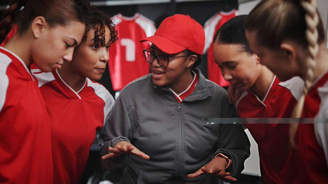 Coach discussing strategy with four young female athletes in red uniforms during a team training session.