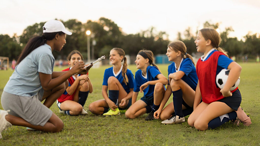 Youth soccer players sit on grass listening to their coach during an outdoor team practice session.