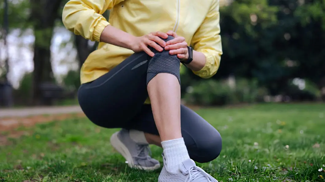 Person in yellow jacket kneeling in a park, holding an injured knee in visible pain.