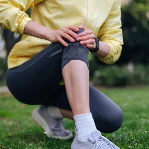 Person in yellow jacket kneeling in a park, holding an injured knee in visible pain.