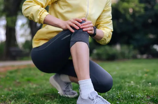 Person in yellow jacket kneeling in a park, holding an injured knee in visible pain.