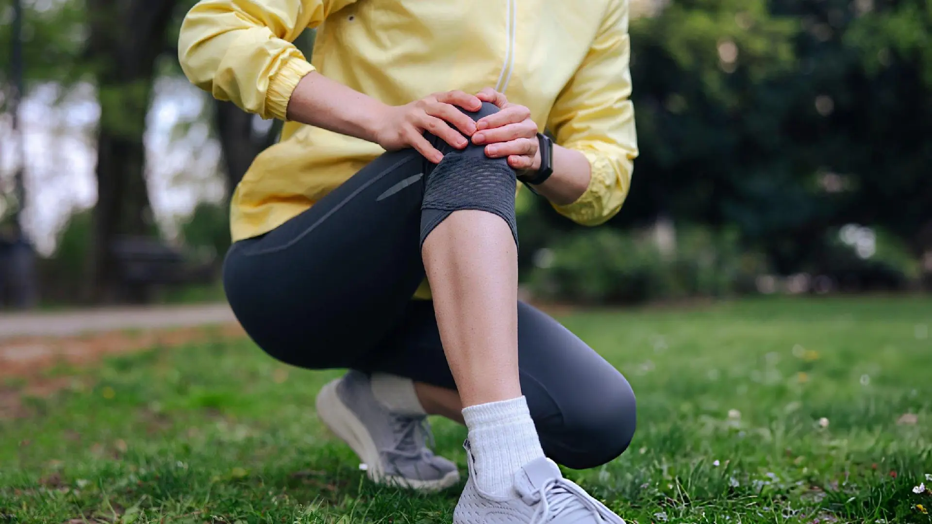 Person in yellow jacket kneeling in a park, holding an injured knee in visible pain.