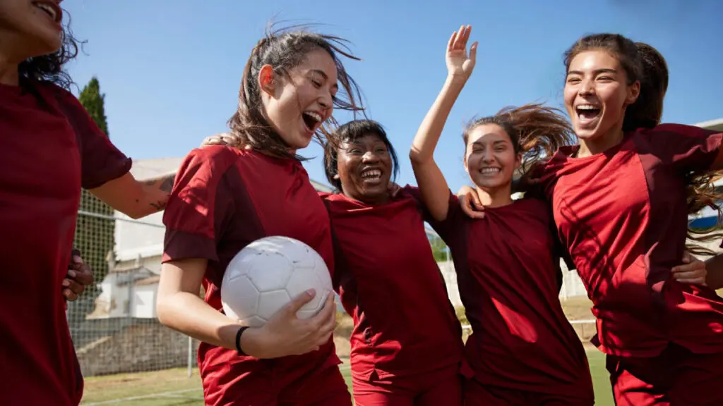 Smiling women's soccer team celebrating victory together, one player holding a white ball outdoors.