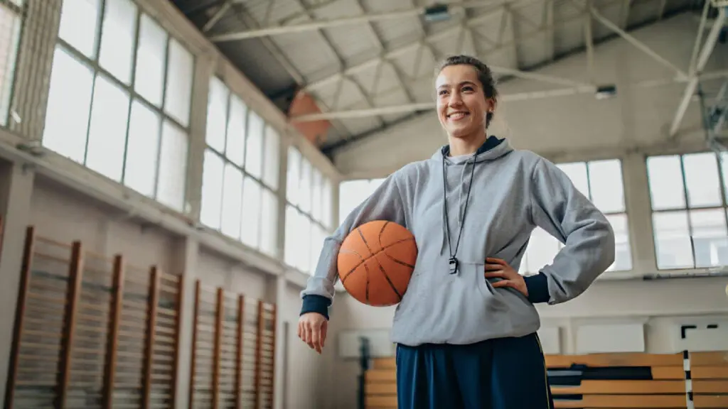Confident female basketball coach smiling in gym holding basketball with whistle around her neck proudly.