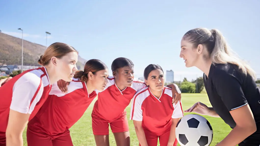 Female soccer coach giving focused instructions to four female players in red jerseys on sunny field.