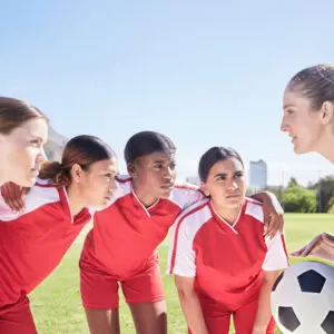 Female soccer coach giving focused instructions to four female players in red jerseys on sunny field.