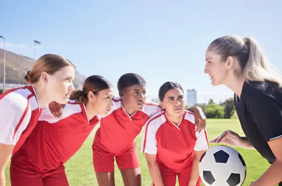 Female soccer coach giving focused instructions to four female players in red jerseys on sunny field.