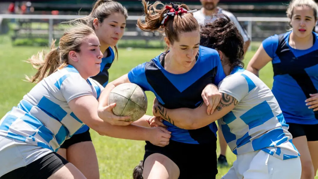 Female rugby player in blue jersey clutches ball while two opponents in striped jerseys tackle her.