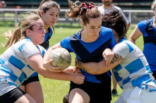 Female rugby player in blue jersey clutches ball while two opponents in striped jerseys tackle her.