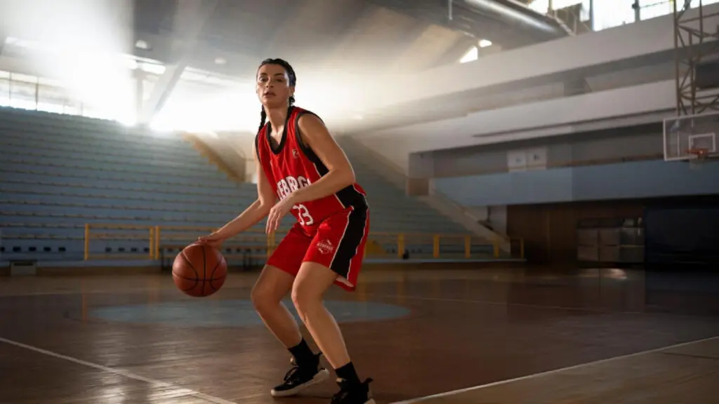 Female basketball player in red jersey dribbles a ball alone in a sunlit empty indoor arena.