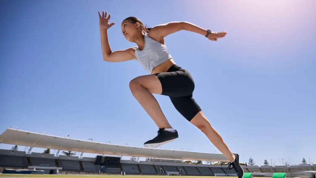 Female sprinter in gray top and black shorts bursts forward on a sunny outdoor stadium track.