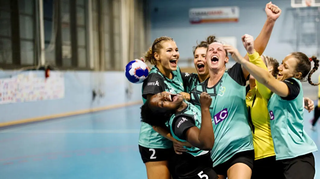 Female handball players in teal jerseys celebrate together, shouting with joy and raising their fists inside an indoor court.