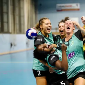 Female handball players in teal jerseys celebrate together, shouting with joy and raising their fists inside an indoor court.