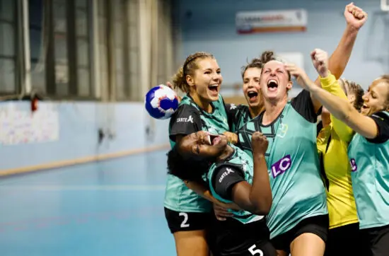 Female handball players in teal jerseys celebrate together, shouting with joy and raising their fists inside an indoor court.