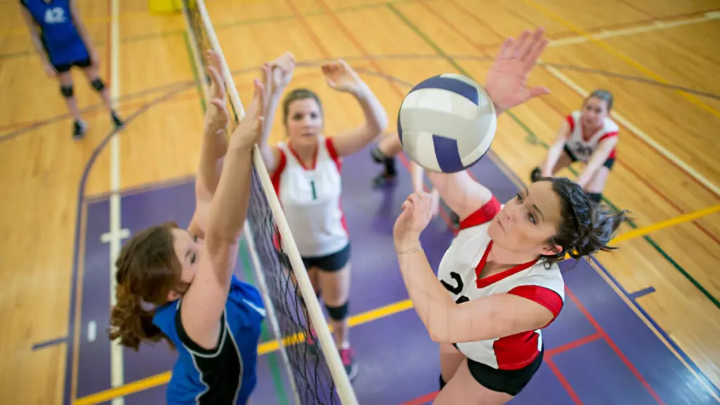 Female volleyball player in red and white jumps to spike the ball while opponents in blue block at net.