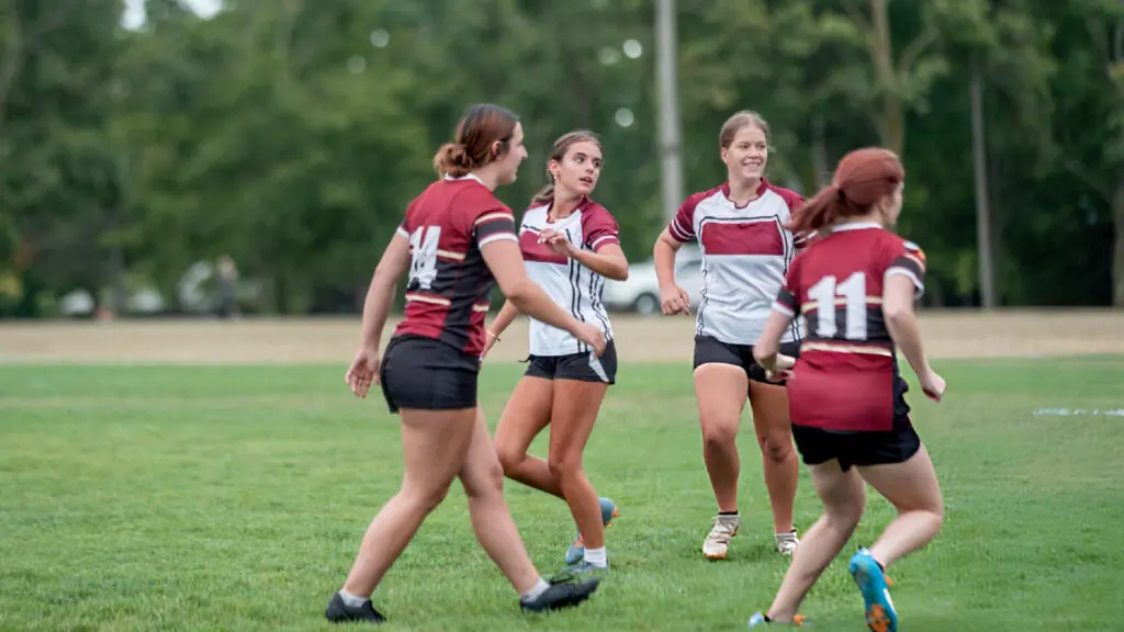 Four female rugby players in maroon and white jerseys run across a green grass field during a match.