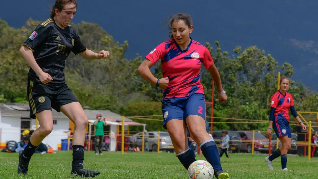 Female soccer player in pink and blue jersey dribbles the ball past a defender in black.