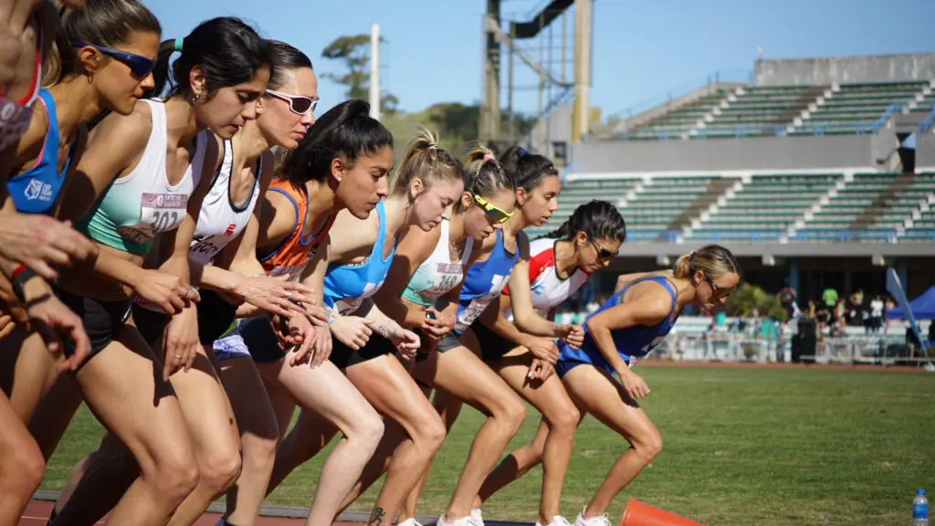 Female runners crouch at the starting line of a track race on a bright sunny day.