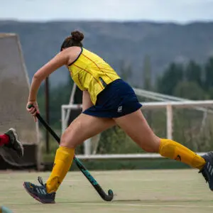 Female field hockey player in yellow uniform sprints with her stick during a competitive match outdoors.