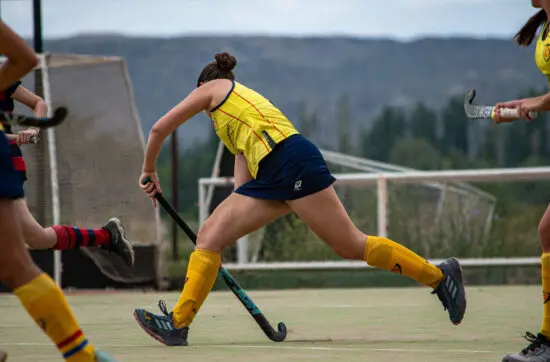 Female field hockey player in yellow uniform sprints with her stick during a competitive match outdoors.