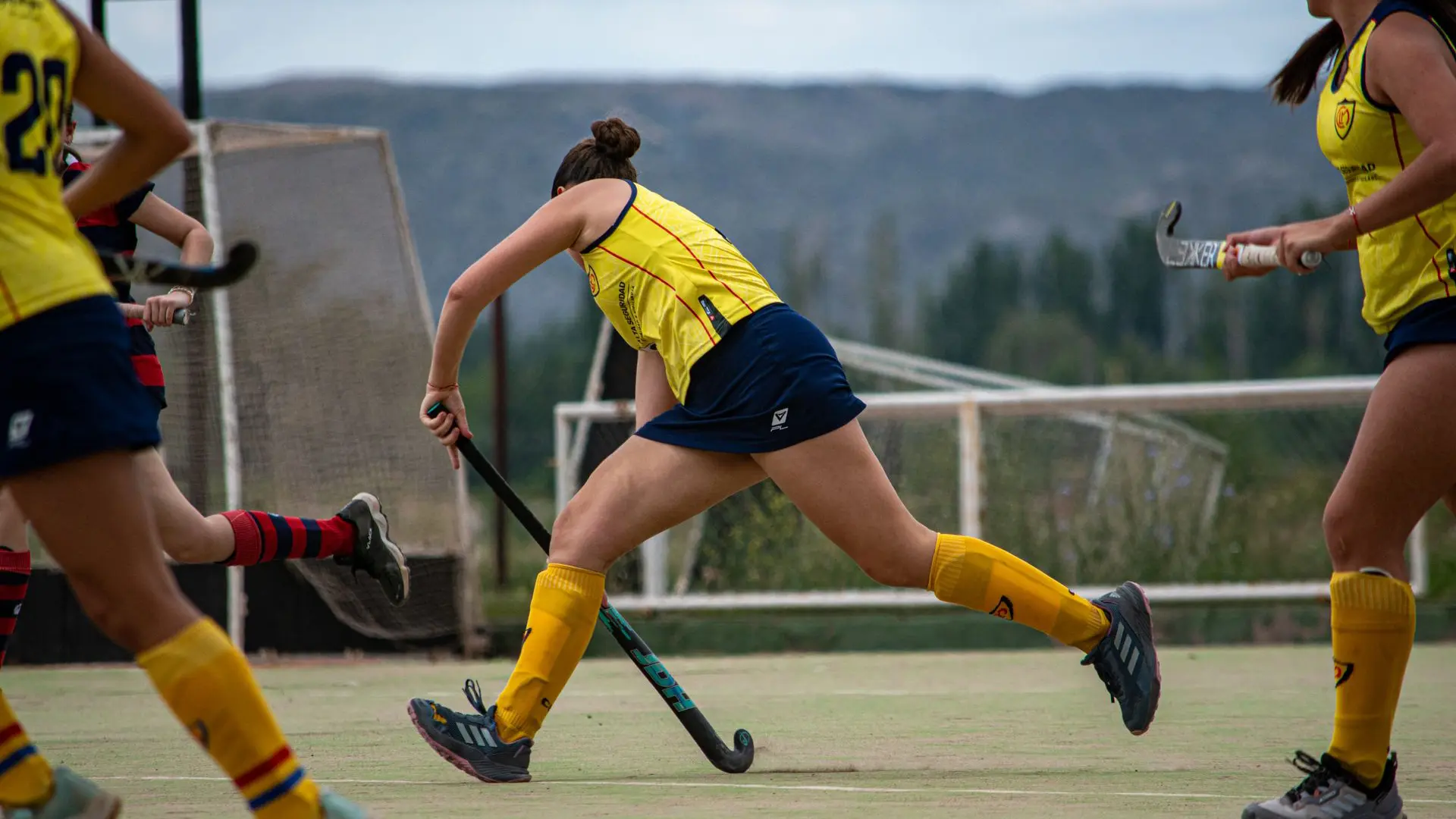 Female field hockey player in yellow uniform sprints with her stick during a competitive match outdoors.